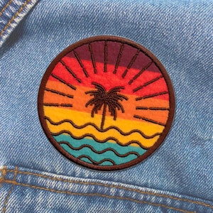 Puede incluir: Un parche redondo bordado con una escena de puesta de sol con una palmera, olas y un arcoíris de colores. El parche es marrón y tiene un contorno blanco.