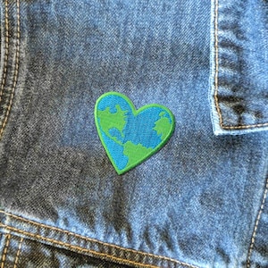 May include: A green and blue embroidered patch in the shape of a heart with a world map design on a blue denim jacket.