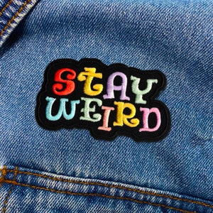 Puede incluir: Parche bordado colorido con las palabras "STAY WEIRD" en tonos arco&iacute;ris sobre un fondo negro. El parche est&aacute; cosido en una chaqueta vaquera azul, mostrando un accesorio divertido y peculiar.
