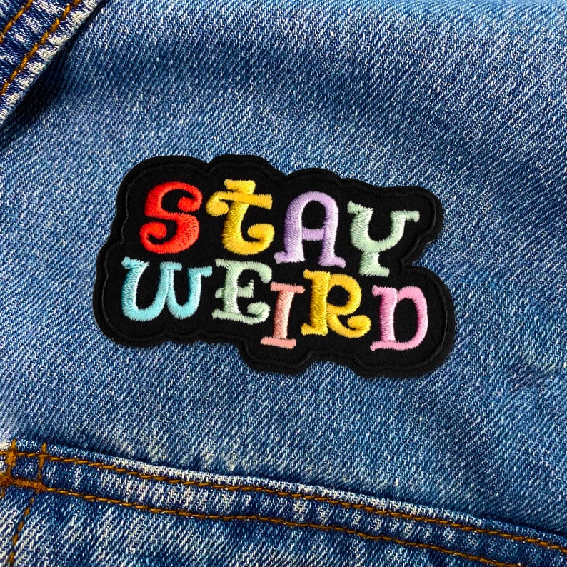 Puede incluir: Parche bordado colorido con las palabras "STAY WEIRD" en tonos arco&iacute;ris sobre un fondo negro. El parche est&aacute; cosido en una chaqueta vaquera azul, mostrando un accesorio divertido y peculiar.