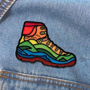 May include: A colorful embroidered patch of a hiking boot. The boot features a vibrant design with layers of red, blue, green, yellow, and orange, resembling a landscape. The patch is outlined in black and has a red sole.