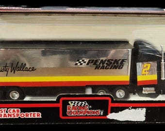 Op de afbeelding: Een model van Rusty Wallace's raceteamtransporter, nummer 2. De zilveren trailer heeft gele en rode strepen en de cabine is zwart. Het model zit in de originele verpakking, met het NASCAR-logo.
