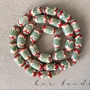 May include: A strand of oval-shaped beads in shades of red, white, and green. The beads have a swirled pattern and are strung together with a gold-colored cord. The text "kas beads" is at the bottom.