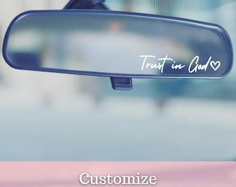 May include: A car's rearview mirror displaying the white script text "Trust in God" with a heart symbol. The mirror is set against a blurred background. The text "Customize the colour" is at the bottom.