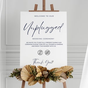 Pode incluir: Um cartaz branco com o texto "Bem-vindos à nossa cerimônia unplugged" e a mensagem "Convidamos você a estar totalmente presente durante nossa cerimônia. Por favor, desligue todos os telefones e câmeras e aproveite este momento conosco." O cartaz também inclui um ícone de proibição de telefone e câmera. O cartaz está em um cavalete com uma decoração de folhas de palma secas e vegetação.