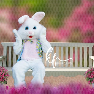 May include: A person in a white Easter bunny costume with blue eyes and a pink nose sits on a white bench. The bunny wears a pink vest and a colorful bow tie. Pink flowers and a blurred background complete the scene.