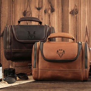 May include: Two leather toiletry bags, one dark brown and one tan, with top handles and zipper closures. The dark brown bag has the initials "M" and "IV" engraved. The tan bag has the name "Harper" and a bow tie engraved.