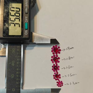 May include: A digital caliper measures a series of small, red, flower-shaped gemstones. The caliper reads 35.68 mm. The gemstones are labeled with sizes ranging from 1.3 mm to 1.9 mm. The caliper is black and silver.