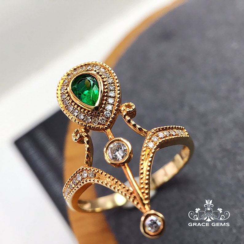 Puede incluir: Un anillo de oro con una esmeralda verde en forma de pera rodeada de peque&ntilde;as piedras preciosas transparentes. El anillo tiene un dise&ntilde;o delicado y ornamentado con detalles intrincados.