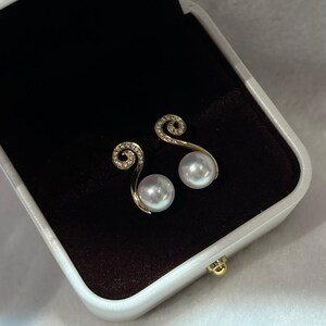 May include: A pair of gold-toned earrings with a pearl and a swirl design. The earrings are presented in a white and black jewelry box. The swirl is encrusted with small, clear stones.