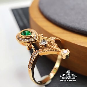 Puede incluir: Un anillo de oro con una piedra preciosa verde en forma de pera rodeada de piedras preciosas transparentes m&aacute;s peque&ntilde;as. El anillo tiene un dise&ntilde;o delicado e intrincado con un motivo floral.