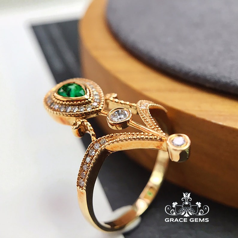 Puede incluir: Un anillo de oro con una piedra preciosa verde en forma de pera rodeada de piedras preciosas transparentes m&aacute;s peque&ntilde;as. El anillo tiene un dise&ntilde;o delicado e intrincado con un motivo floral.