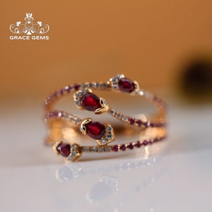 May include: A gold ring with a delicate design featuring four red gemstones and small white diamonds. The ring is set on a white surface.