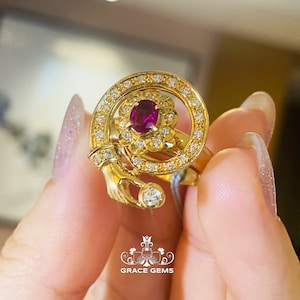 May include: A gold ring featuring a central oval ruby surrounded by a halo of small diamonds. The ring has a unique design with a swirling pattern and additional diamond accents. The ring is from Grace Gems.
