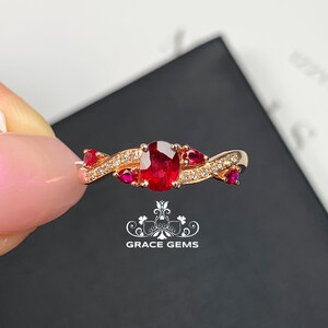 May include: Rose gold ring featuring an oval-cut ruby center stone, flanked by smaller rubies and diamond accents. The band has a twisted design. The ring is held against a black background.
