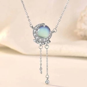 May include: A silver necklace with a crescent moon pendant featuring a round iridescent gemstone and a row of small clear stones. The pendant has two dangling chains with a star charm at the end of each chain.