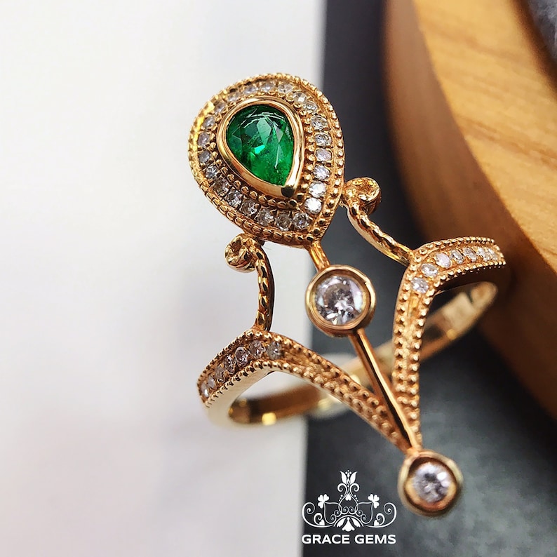 Puede incluir: Un anillo de oro con una esmeralda en forma de pera rodeada de diamantes m&aacute;s peque&ntilde;os. El anillo tiene un dise&ntilde;o delicado e intrincado con una parte superior puntiaguda en forma de corona.