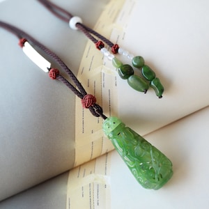 Może przedstawiać: Naszyjnik z zielonym jadeitowym wisiorem o skomplikowanych rzeźbach. Wisiorek jest przymocowany do brązowego sznurka ze srebrną tabliczką z imieniem i zielonymi koralikami z jadeitu.