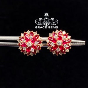 Puede incluir: Un par de pendientes de oro con un racimo de rubíes rojos y diamantes blancos en forma de flor. Los pendientes están sobre un fondo plateado.
