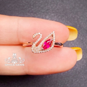 Può includere: Un anello in oro rosa con un design a forma di cigno adornato con una pietra preziosa di rubino rosso al centro. Il cigno è decorato con piccole pietre preziose trasparenti.