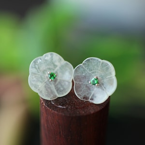 May include: A pair of white flower-shaped earrings with green gemstone accents. The earrings are displayed on a wooden surface.