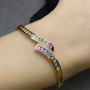 May include: A gold-toned hinged bangle bracelet featuring a bypass design with channel-set square-cut gemstones in a rainbow gradient. The bracelet includes rows of small pavé-set clear stones along the center overlapping section.