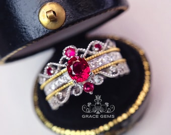 May include: A silver ring featuring a large red ruby gemstone, surrounded by smaller diamonds and a delicate filigree design. The ring is displayed in a black ring box with gold trim.