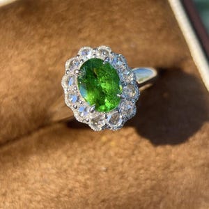 May include: A silver ring featuring a large, oval-cut, vibrant green gemstone at the center. The gemstone is surrounded by a halo of smaller, clear gemstones, creating a sparkling, floral-like design. The ring is set against a brown background.