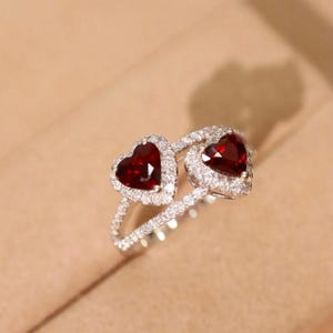 Pode incluir: Um anel de prata com duas pedras preciosas vermelhas em forma de coração, cercadas por pedras brancas menores.