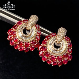 May include: A pair of gold earrings with a circular design featuring a halo of small diamonds and a larger center diamond. The earrings are accented with a cluster of red rubies.