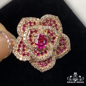 May include: A rose-shaped ring featuring a large red ruby gemstone surrounded by smaller red rubies and white diamonds set in gold.