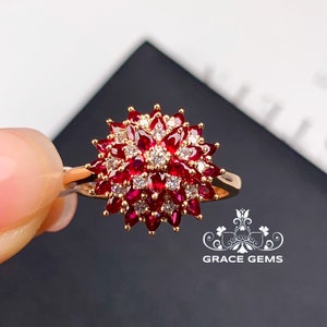 May include: A gold ring featuring a large red ruby and smaller diamonds set in a flower-shaped design. The ring is on a finger.
