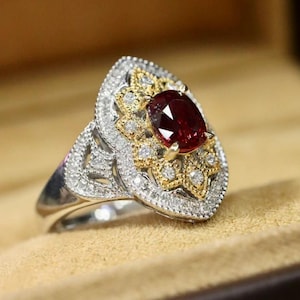 May include: An ornate ring featuring a large, oval-cut ruby gemstone. The ring has a silver band and a detailed setting with gold accents and small diamonds. The design is intricate and elegant, showcasing a vintage or antique style.