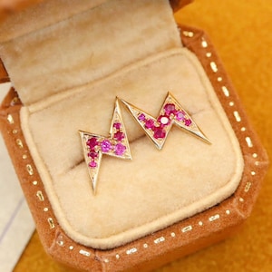 May include: A pair of gold lightning bolt earrings with pink gemstones. The earrings are in a small brown velvet box.