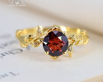 May include: A gold ring with a red gemstone set in a leafy design. The ring is on a white surface.