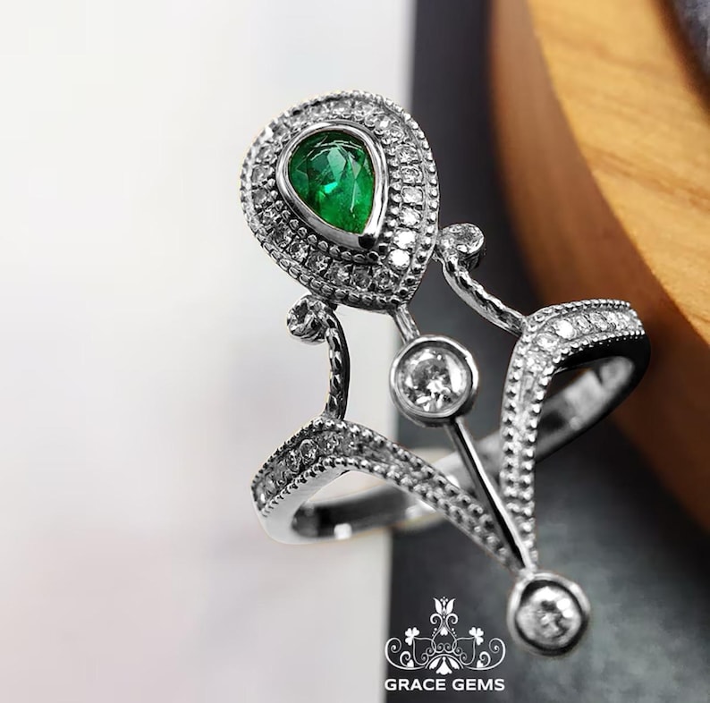 Puede incluir: Un anillo de plata con una esmeralda en forma de pera rodeada de diamantes m&aacute;s peque&ntilde;os. El anillo tiene un dise&ntilde;o delicado e intrincado con una forma de corona.