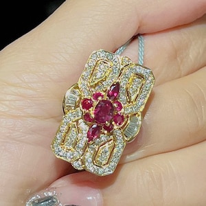 Puede incluir: Un anillo de oro con un gran rubí rojo rodeado de diamantes más pequeños. El anillo está engastado en una forma rectangular con detalles intrincados.