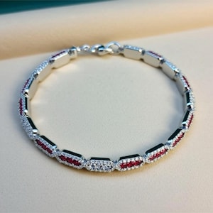May include: A silver bracelet with alternating sections of red gemstones and clear crystals. The bracelet is linked together with a clasp. The gemstones and crystals are set in a silver metal.