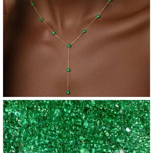 May include: A delicate gold necklace featuring emerald green gemstones. The necklace has a Y-shaped design with multiple emeralds along the chain and a single emerald at the bottom. The image also shows a pile of loose emeralds.