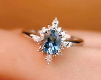 May include: A silver ring featuring an oval, light blue gemstone surrounded by smaller, clear gemstones. The ring's design is a halo style, with the smaller stones arranged around the central stone, creating a sparkling effect. The ring is set against a warm, neutral background.