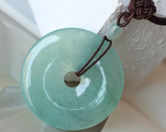 May include: A light green jade pendant in the shape of a circle with a hole in the center. The pendant is hanging from a brown cord.
