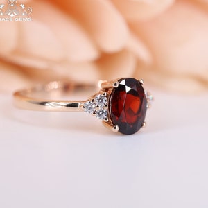 May include: A rose gold ring with a large oval-shaped garnet gemstone surrounded by smaller round diamonds. The ring is set on a white background.