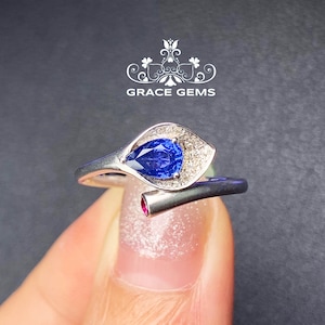 May include: A pear-shaped blue sapphire ring set in a white gold band with a delicate floral design. The ring features a small ruby accent on the side.