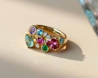 Könnte beinhalten: Ein goldener Ring mit einer Ansammlung farbenfroher Edelsteine. Der Ring zeigt eine Anordnung von Edelsteinen in verschiedenen Formen und Größen, darunter Blau, Rosa, Grün und Lila, auf einem goldenen Band. Die Edelsteine sind in einem dekorativen Muster angeordnet.