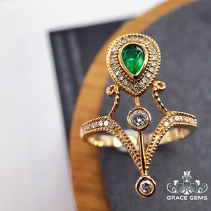 Puede incluir: Un anillo de oro con una esmeralda verde en forma de pera rodeada de diamantes m&aacute;s peque&ntilde;os. El anillo tiene un dise&ntilde;o delicado e intrincado con una parte superior puntiaguda en forma de corona y un diamante colgante en la parte inferior.