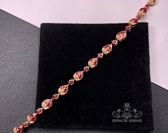 May include: A gold bracelet with teardrop-shaped ruby and diamond accents. The bracelet is on a black velvet background.  GRACE GEMS is printed on the background.