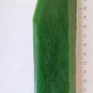 Peut inclure: Une pierre de jade rectangulaire polie, vert foncé. Une règle est placée à côté de la pierre pour l'échelle, montrant une longueur d'environ 23 cm. Le jade a une surface lisse et réfléchissante avec des variations naturelles visibles.