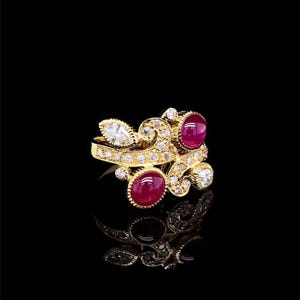 Può includere: Un anello d'oro dal design dettagliato, con due pietre rosse taglio ovale e diverse piccole pietre trasparenti. L'anello ha uno stile decorativo e ornato con una superficie riflettente.