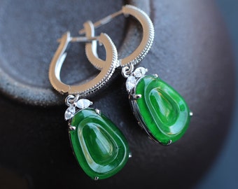 May include: A pair of silver hoop earrings with a teardrop-shaped jade stone pendant. Each pendant is accented with small, clear gemstones.