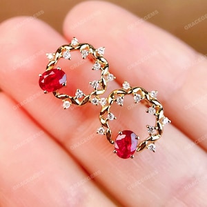 Puede incluir: Un par de pendientes de oro con un diseño delicado y trenzado. Cada pendiente presenta una piedra preciosa de rubí rojo engastada en el centro del diseño.